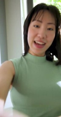 Dikey video: Yirmili yaşlarda Çinli bir kadın içeri giriyor, kamerayı selamlıyor, bitkilerin yanında atıştırmalıkları tanıtıyor. Dost canlısı, hayat dolu, rahat, doğal, nişanlı, dikey video