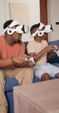 Vertical video: African American dad and son donning VR headsets at home playing and cheering. Immersive, interactive, digital, entertainment, dynamic, family, vertical video