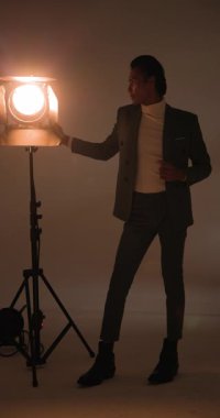 Dikey video: ceketli Asyalı erkek sahne ışıkları altında stüdyoda çekim için ahır kapılarını ayarlıyor. Profesyonel, moda, minimalist, ışıklandırma, erkek, portre, dikey video