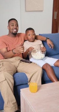 Dikey video: Afrikalı Amerikalı baba ve oğul televizyona tepki veriyor, gülüyor, kanalı değiştiriyor. Aile, bağ, rahatlama, eğlence, konfor, rahatlık, dikey video