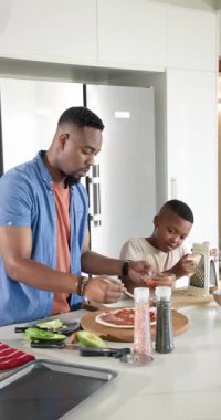 Dikey video: Afrika kökenli Amerikalı baba ve oğul avokado dilimliyor, evde pizza için peynir rendeliyorlar. Aile, kaynaşma, yemek pişirme, beslenme, ev, sağlıklı, dikey video