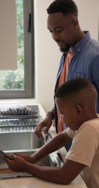 Dikey video: Afro-Amerikan baba mutfağa giriyor, oğluna musluk altında bulaşık yıkamayı öğretiyor. Aile, kaynaşma, eğitim, yerli, işçilik, ev işi, dikey video