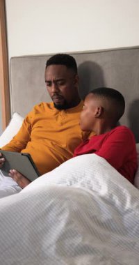Dikey video: Afrika kökenli Amerikalı baba ve oğul yatak odasında sesli bir şekilde kitap okuyor. Ebeveynlik, bağlanma, eğitim, hikaye anlatma, samimi, samimi, dikey video