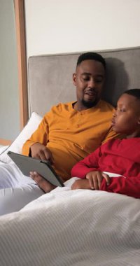Dikey video: Afro-Amerikan baba yatakta tablet kullanıyor ve oğlunu yorganın içine sokuyor. Aile, bağlanma, yetiştirme, eğitim, yatma zamanı, konfor, dikey video