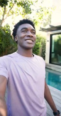 Dikey video: Arka bahçede yürürken Afrikalı Amerikalı bir adamın havuz kenarında çekim yapmasını selamlıyorum. Açık hava, yaşam tarzı, modern, boş zaman, vlogger, güneş ışığı, dikey video
