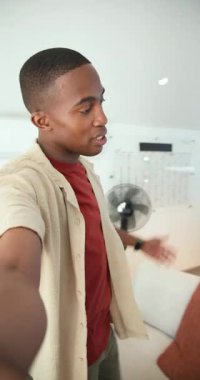Dikey video: Afro-Amerikalı adam telefon sallıyor, pansiyon odası, kanepede vlog 'a oturuyor. Teknoloji meraklısı, modern iç mekan, içerik oluşturma, yaşam tarzı, ev ofisi, günlük tarz, dikey video