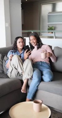 Dikey video: Kanepede ekranları paylaşmadan önce bayan arkadaşlar akıllı telefonları kontrol ediyorlar. Arkadaşlık, rahatlama, rahatlama, modern, rahat, teknoloji, dikey video