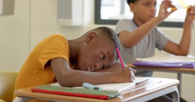 Okulda, Afrikalı Amerikalı bir çocuk masa başında dinleniyor, elinde kalem, düşünceli görünüyor. Eğitim, öğrenci, sınıf, öğrenme, çalışma, tefekkür