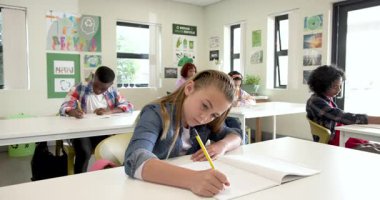 Sınıfta, okulda odaklanmış sınıf arkadaşlarıyla birlikte not defterine yazan gülümseyen kız. Eğitim, okul, öğrenim, öğrenim, çok ırklı öğrenciler, konsantrasyon