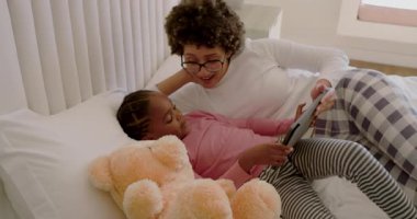 Çeşitli anne ve kız tablet tıkırtılarına yanıt veriyor, beyaz yatağa dokunuyor, hikaye kitabının sayfalarında geziniyor. Aile, bağ, rahatlama, rahatlık, dijital öğrenme, yetiştirme, ev ortamı