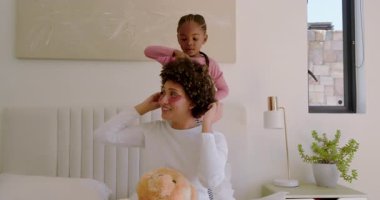 Afro-Amerikalı kız, annesinin oyuncak ayıyı yatakta tuttuğunu fark ettikten sonra saçlarını pembe tarıyor. Aile, bağlar, bakım, şefkat, saç bakımı, şefkat, ev