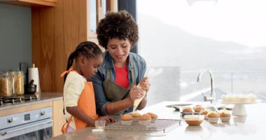 Anne, kızının mutfak adasında keklerin üzerine şekerleme dökmesine rehberlik ediyor, kırmızı şeker serpiyor. Aile, pişirme, aşçılık, mutfak, zanaat, yaratıcılık, etkinlik