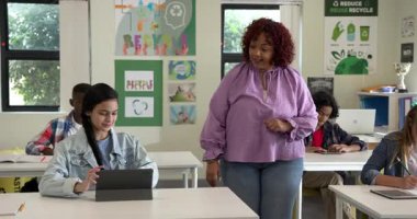 Sınıfta tabletli bir kıza yardım eden bayan öğretmen, okulda interaktif öğrenmeyi teşvik ediyor. Eğitim, teknoloji, dijital, öğretim, akıl hocalığı, işbirliği