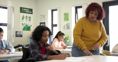 Tekerlekli sandalyedeki çocuğa sınıfta yazma konusunda yardım eden bayan öğretmen, okulda öğrenmeyi teşvik ediyor. Eğitim, çeşitlilik, kapsama, öğretim, yardım, destek