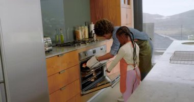 Afro-Amerikan anne ve kızı mutfakta fırın saatinin sesini duyuyorlar, çörekleri soğutma rafına kaydırıyorlar. Aile, pişirme, aşçılık, ev, gelenek, sıcaklık, birliktelik.