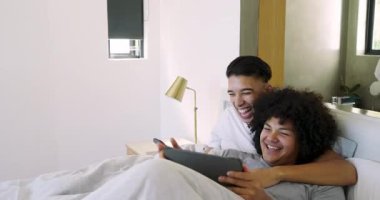 Yatak odasındaki yastıklarla yatağa uzanan erkek partnerler tablet kaydırıyor ve akıllı telefonu paylaşıyorlar. Yakınlık, rahatlama, bağlanma, teknoloji, rahatlık, sıcaklık.