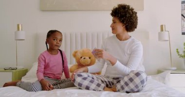 Afro-Amerikalı anne yatakta paketi açıyor, kızı göz bandı altında başvuruyor. Aile, cilt bakımı, kaynaşma, rahatlama, rahatlık, samimiyet, birliktelik.