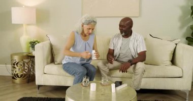 Üst düzey çift oturma odasına ellerinde su bardaklarıyla girip sağlık için hap yutuyor. İlaç, sağlık, sağlık, destek, yaşam tarzı, konfor, rahatlama