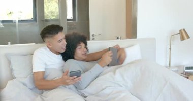 Yatakta oturmuş akıllı telefon ekranını işaret eden iki adam tableti kapıyor, içeriklerini gösteriyor ve gülüyorlar. Samimiyet, şefkat, bağlantı, rahatlama, modern, samimi, boş zaman