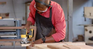 Kırmızı tişört ve gözlük takan Afro-Amerikan bir adamı yapboz atölyesinde kalas keserek çalıştırmak. Zanaatkar, marangozluk, marangozluk, atölye, aletler, işçilik