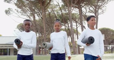 Çok ırklı öğrenciler yoga minderlerini sahada tutuyorlar, gülümsüyorlar ve okul etkinliğine hazırlar. Çok ırklı öğrenciler, yoga pratiği, egzersiz, gülümseme, spor, eğlence.