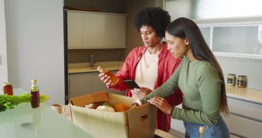 Cam Ada 'da market kutularını açan, telefondaki eşyaları doğrulayan, tezgaha yerleştiren çeşitli çiftler. Taze ürün, modern mutfak, sağlıklı yaşam tarzı, aşçılık, yemek hazırlama, ev yapımı, canlı.