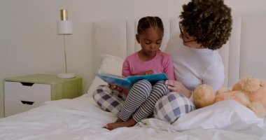 Anne ve kızı çocuk gibi kaynaşıyor mavi resim kitabını açıyor, kitap okuyor ve yatakta kıkırdıyor. Aile, bağ, eğitim, hikaye anlatma, samimiyet, çocukluk, yetiştirme