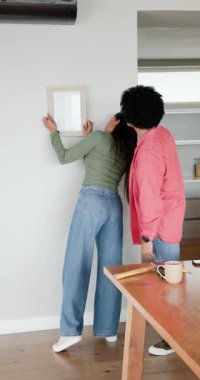 Dikey video: Hintli bir çift el kol hareketi yapıyor, evdeki duvara çerçeve asıyor ve memnuniyetle sarılıyorlar. Romantik, işbirliği, ev, iç mekan, bağ, samimi, dikey video