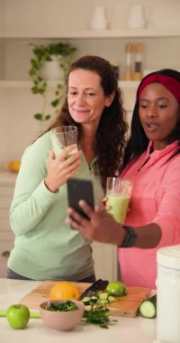 Dikey video: Çeşitli bayan arkadaşlar telefonda selfie pozu veriyor ve mutfakta smoothie yudumluyor. Arkadaşlar, dostluk, sağlık, yaşam tarzı, sağlıklı, dijital, dikey video