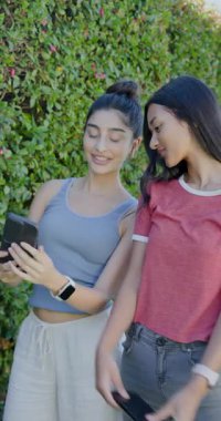Çeşitli kadın arkadaşlar telefon ve ekran sunuyor, selfie çekiyor ve bahçede kayıyor. Arkadaşlık, bağlantı, açık hava, canlı, yaşam tarzı, iletişim, dikey video