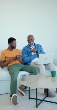 Dikey video: Afro-Amerikalı baba-oğul telefon ekranını paylaşıyor ve ev kanepesinde dizüstü bilgisayar kullanıyor. Aile, danışmanlık, bağlantı, teknoloji, rahatlama, iletişim, dikey video