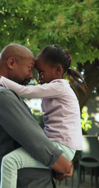 Dikey video: Afrikalı Amerikalı baba-kız kucaklaşması sonra da onu arka bahçeye bağlamak için kaldırması. Aile, bağ, sevgi, destek, açık hava, doğa, dikey video