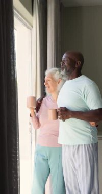 Dikey video: Sürgülü camdan içeri güneş girerken kupalarla kucaklaşan yaşlı çift. Şefkat, arkadaşlık, yaşam tarzı, konfor, dinginlik, sıcaklık, dikey video