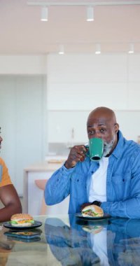 Dikey video: Baba bardak kaldırıyor, oğul mutfakta bardak tokuşturuyor ve kahvaltıda telefona bakıyor. Aile, yakınlaşma, sohbet, boş zaman, ev, arkadaşlık, dikey video