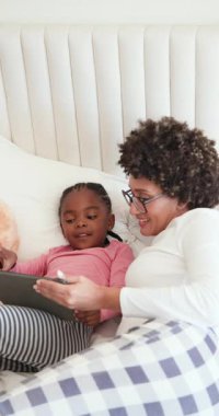 Afrika kökenli Amerikalı anne, yastıkların ve ayıların arasında yatağın üzerinde uygulamaları keşfetmek için tablet tıklatan kızına rehberlik ediyor. Aile, bağ, öğrenme, eğitim, teknoloji, ev, dikey video