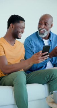 Dikey video: Afro-Amerikan baba-oğul uygulamalar kullanarak evde telefonları dinliyorlar ve gülüyorlar. Teknoloji, akıl hocalığı, iletişim, aile içi, rahat, rahat, dikey video