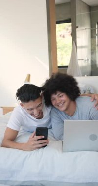 Akıllı telefonu kaldırıyor, iki erkek arkadaş yatağında videoları aşırıyor, keyfi paylaşmak için, dizüstü bilgisayara doğru eğiliyor. Arkadaşlık, rahatlama, rahatlama, konforlu, bağlanma, iletişim, dikey video