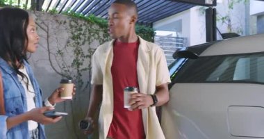 Eve elinde kahveyle gelen Afro-Amerikan çift EV kablosunu kesiyor ve birbirlerine gülümsüyor. Yoldaşlık, kentsel, canlı, modern, yaşam tarzı, gündelik, teknoloji