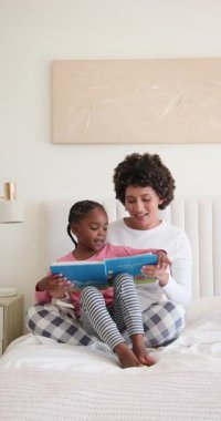 Annem hikaye saati için renkli resim kitabı açıyor ve yatak odasında anaokulu kızına yüksek sesle okuyor. Aile, bağ, çocukluk, eğitim, konforlu, besleyici, dikey video