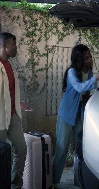 Ev yoluna yaklaşan Afro-Amerikan çift bagaja bavul dolduruyor ve telefonları karşılaştırıyor. Dost canlısı, seyahat, macera, yaşam tarzı, günlük, açık hava, dikey video