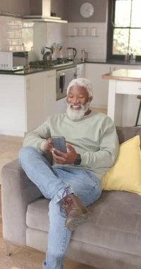 Dikey video: Son sınıf Afrikalı Amerikalı adam koltuğa yaslanmış, akıllı telefonuyla mutfakta gülümsüyor. Günlük, rahatlama, modern, teknoloji, konfor, yaşam tarzı, dikey video