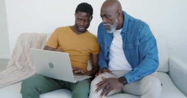 Afrika kökenli Amerikalı baba ve oğul, ders çalışmayı ve gülmeyi öğrenmek için kanepede dizüstü bilgisayarla iş birliği yapıyorlar. İşbirliği, akıl hocalığı, nesiller arası, takım çalışması, rahat, rahat, elektronik