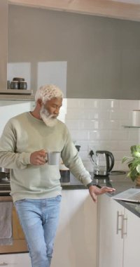 Dikey video: Yetişkin bir Afro-Amerikan adam mutfakta dizüstü bilgisayara bakıyor ve bardak tutuyor. Ev, yaşam tarzı, çağdaş, teknoloji, rahatlama, gündelik, dikey video