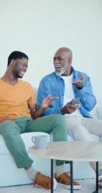 Dikey video: Afro-Amerikalı baba ve oğlu kanepede kahkahalarla kahkahalar atıyorlar. Aile, akıl hocalığı, teknoloji, öğrenme, bağlanma, konfor, dikey video