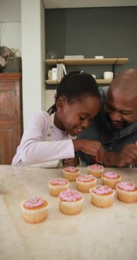 Dikey video: Afrika kökenli Amerikalı bir kız kekleri kepçe ile kapkeklerin üzerine serpiyor ve baba evde rehberlik ediyor. Aile, bağ, yaratıcılık, yerli, sıcaklık, kutlama, dikey video