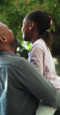 Dikey video: Gülen Afrikalı Amerikalı baba kızını arka bahçede ağacın altına kaldırıyor, çene ovuyor. Aile, bağ, açık hava, doğa, sevgi, oyunculuk, dikey video