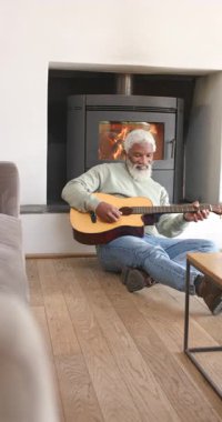 Dikey video: Gitar taşıyan, soba tıngırdatan yaşlı Afrikalı Amerikalı adam. Rahat, rahatlatıcı, akustik, ev, konfor, sıcak, dikey video