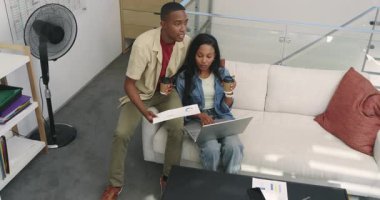 Afro-Amerikalı çift sehpanın üzerinde kayıyor ve kahve yudumlayıp dizüstü bilgisayarın verilerini inceliyor. İşbirliği, ortaklık, çalışma alanı, gündelik, modern, iletişim, üretkenlik