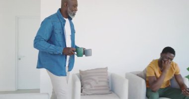 Kıdemli Afro-Amerikan babası oturma odasına elinde fincanlarla giriyor. Akıllı telefon görüntüsünden sonra oğlunu teselli ediyor. Aile, bağ, rahatlama, rahatlık, iç mekan, sıcaklık, destek