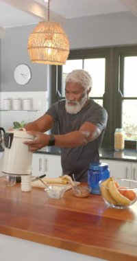 Dikey video: Smoothie Senior Man kaldırıyor blender açıyor mutfakta muz dilimliyor. Modern, aşçı, sağlıklı, canlı, besleyici, evcil, dikey video